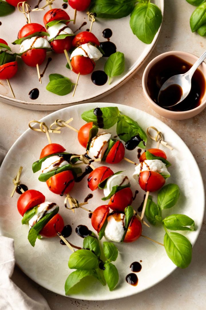 two plates served with caprese skewers garnished with blasamic glazed and basil leaves. A small bowl with a spoon and more balsamic glaze on the side