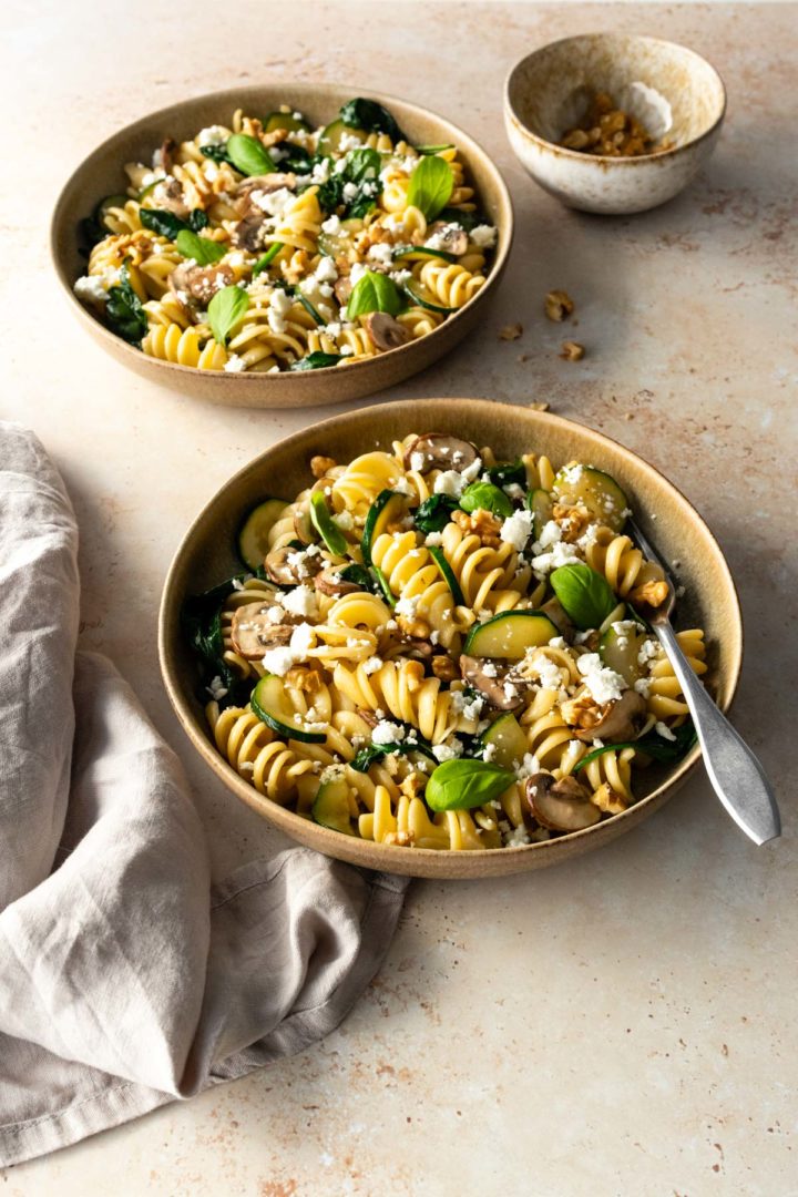 two plates with pasta, sliced zucchini, walnuts, crumbled feta, mushrooms, spinach and fresh basil. One small bowl with walnuts on the side