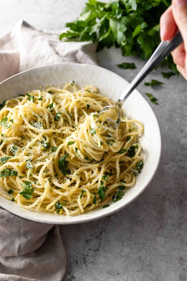 a bowl with spaghetti pasta with lemon zest, parmesan and chopped parsley. More parsley on top of the board and a fork with more pasta