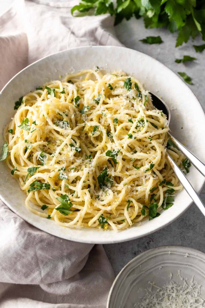a large bowl with lemon pasta, a small bowl with grated parmesan and parsley on top of a board