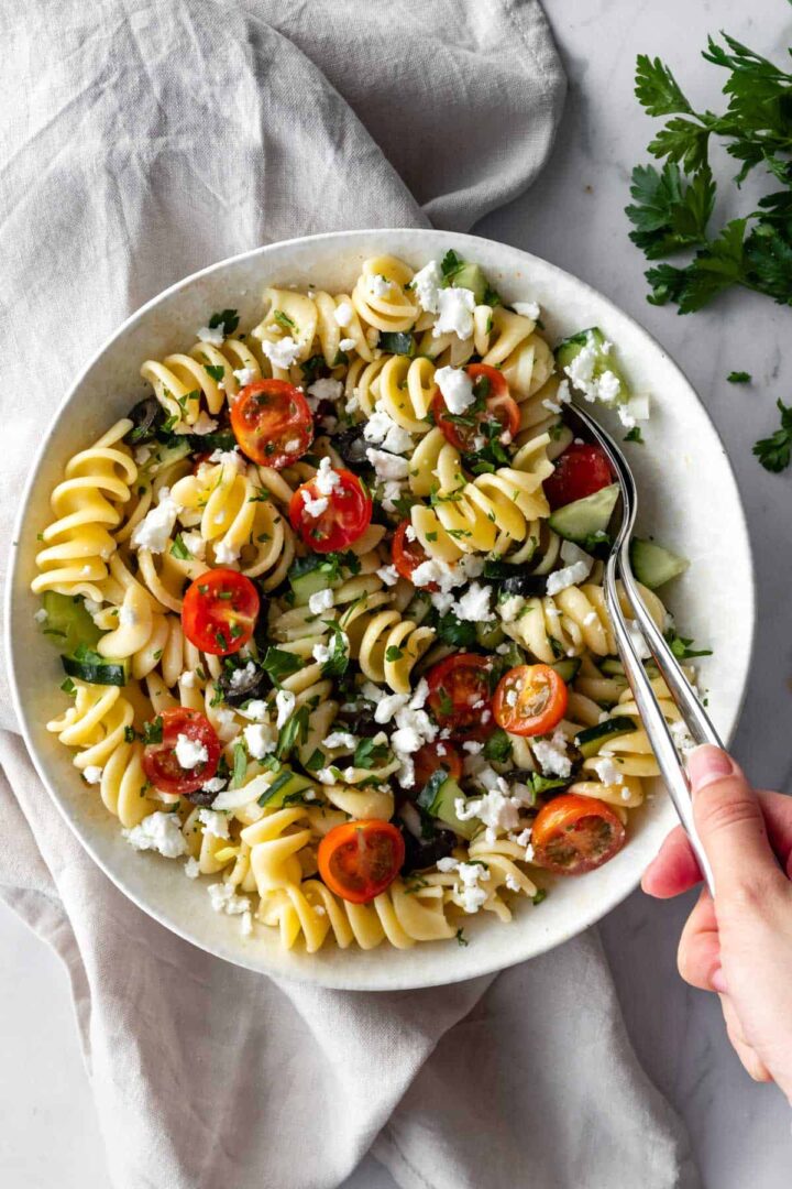 a bowl with a pasta salad with cherry tomatoes, olives, cucumber, parsley and crumbled feta cheese. One hand holding serving spoons