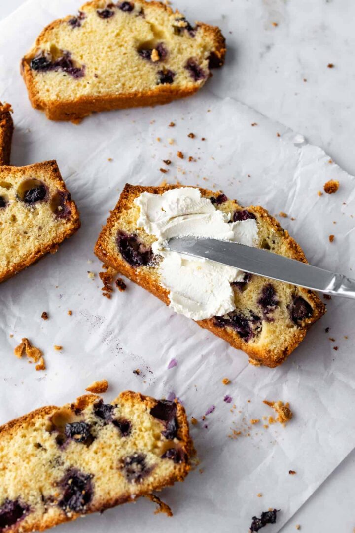 slices of lemon loaf with blueberries, a knife on one of them with cream cheese