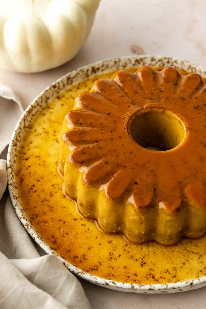 a pumpkin spice flan served on a large plate, a white pumpkin on the top left corner, a kitchen linen towel on the bottom left corner
