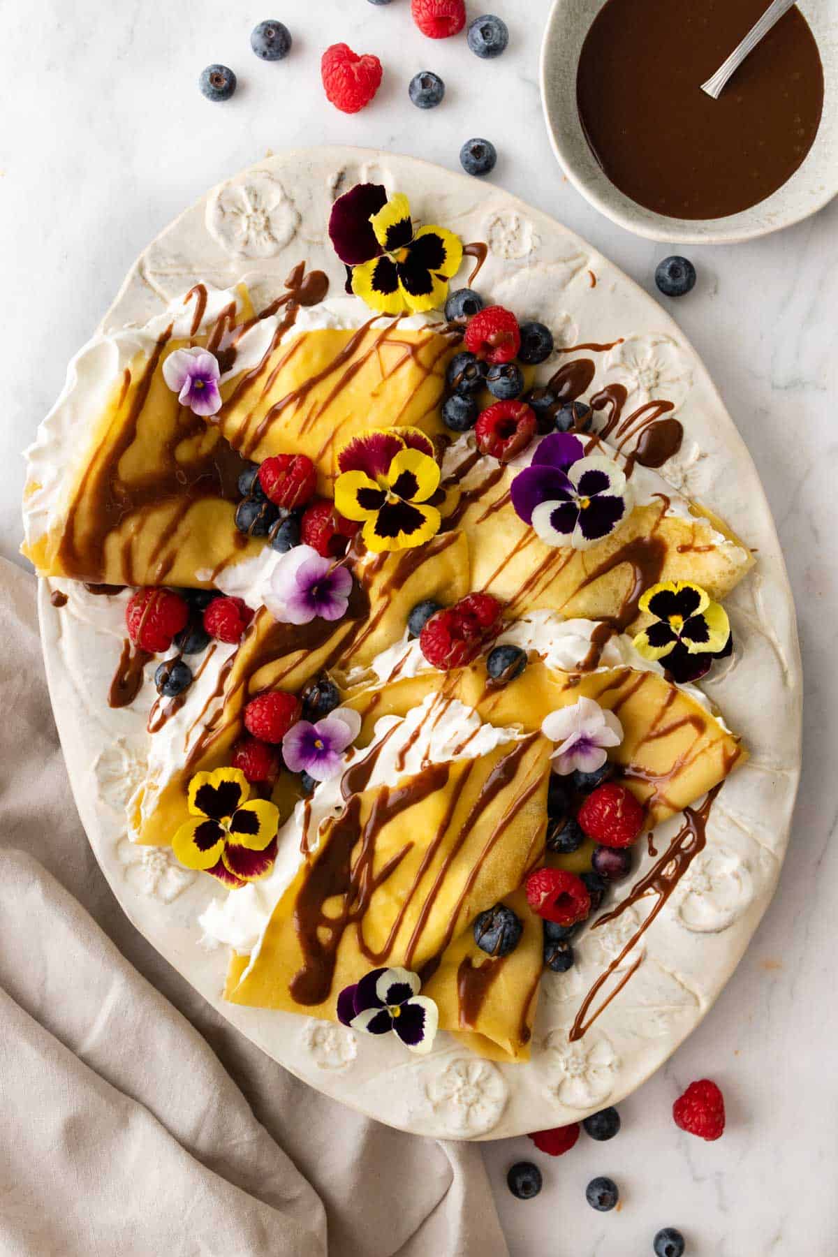 a large plate with crepes and whipped cream inside the crepes. The crepes are garnished with berries, chocolate sauce, and flowers. More berries, a kitchen towel, and a small bowl with more chocolate sauce on the side.