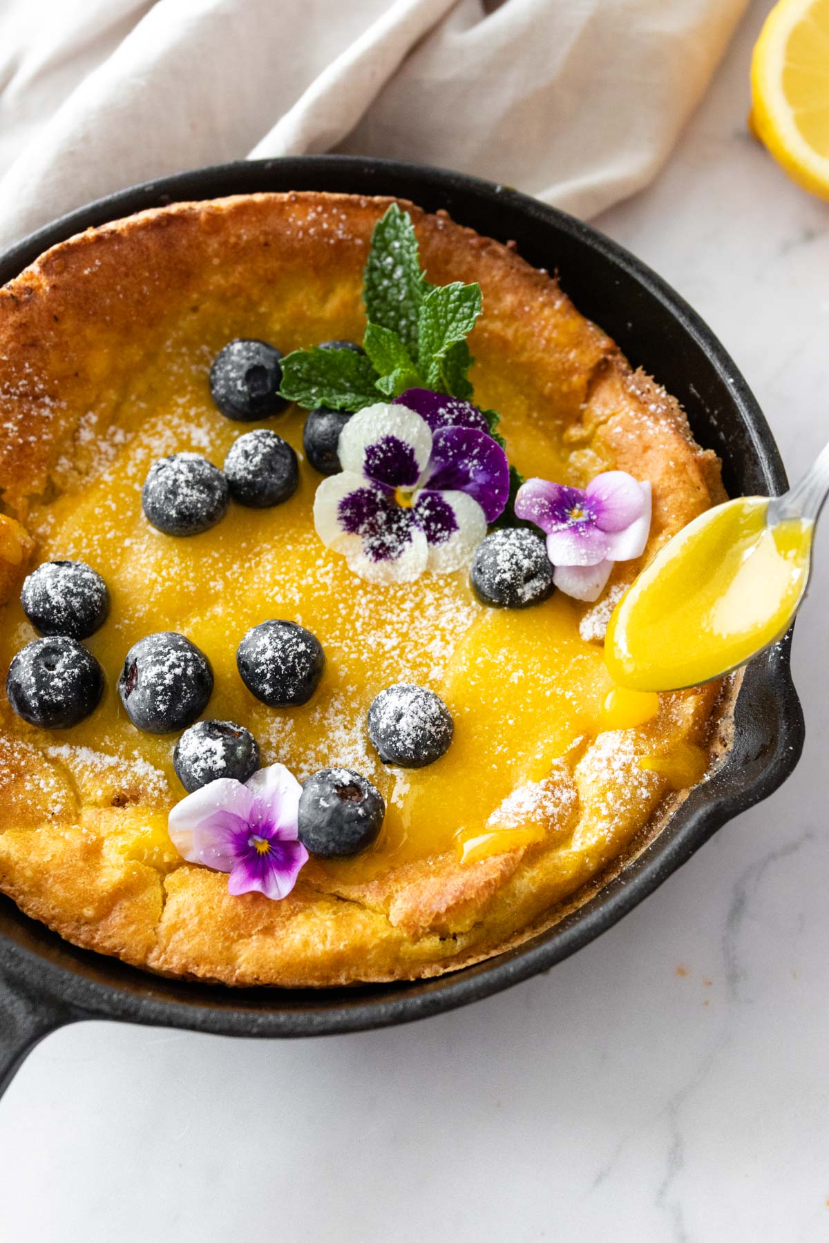 a iron skillet with a dutch baby pancake with lemon curd, blueberries, mint, edible flowers, and powdered sugar. A spoon with lemon curd on the side.